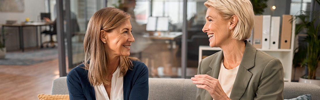 Two female employees engaged in a deep conversation on a communal workplace sofa, showing employee wellbeing support in action