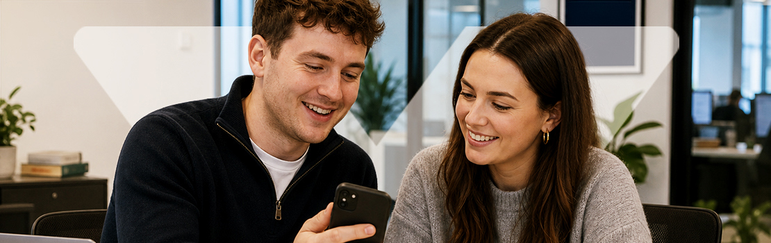 Two smiling employees in a modern office space looking at employee discounts on a phone, showing the value of why companies offer employee discounts